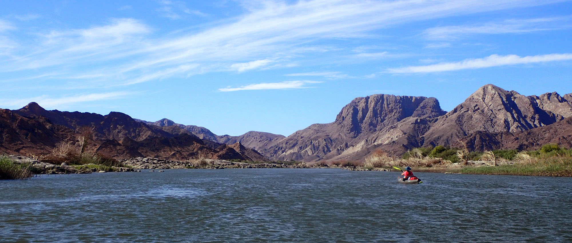 A lone kayaker paddling a wide stretch of the Orange River through arid mountains.
